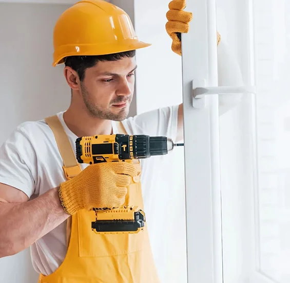 Handyman in yellow hard hat and overalls using a cordless power drill to install or repair a window frame in a home.