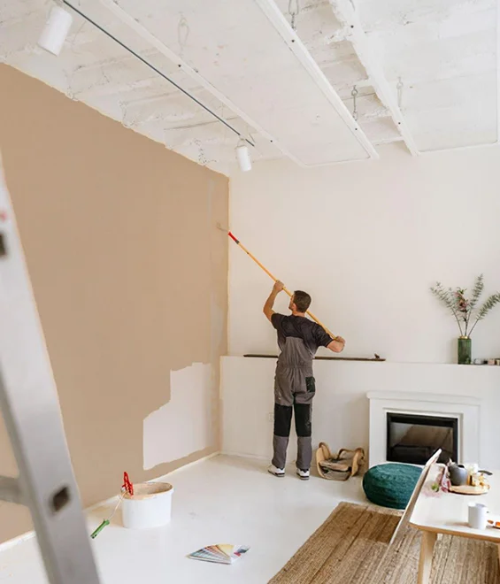 Person painting a white wall in a modern living room during a home remodeling project, with ladder and renovation tools in view.